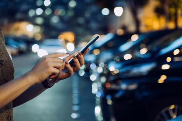 Billedet viser en kvinder, der holder hendes mobiltelefon. Du kan ikke se andet end kvindens hænder, da fokusset er på mobiltelefonen. Hun er placeret på en parkeringsplads, hvor der er en masse biler parkeret.