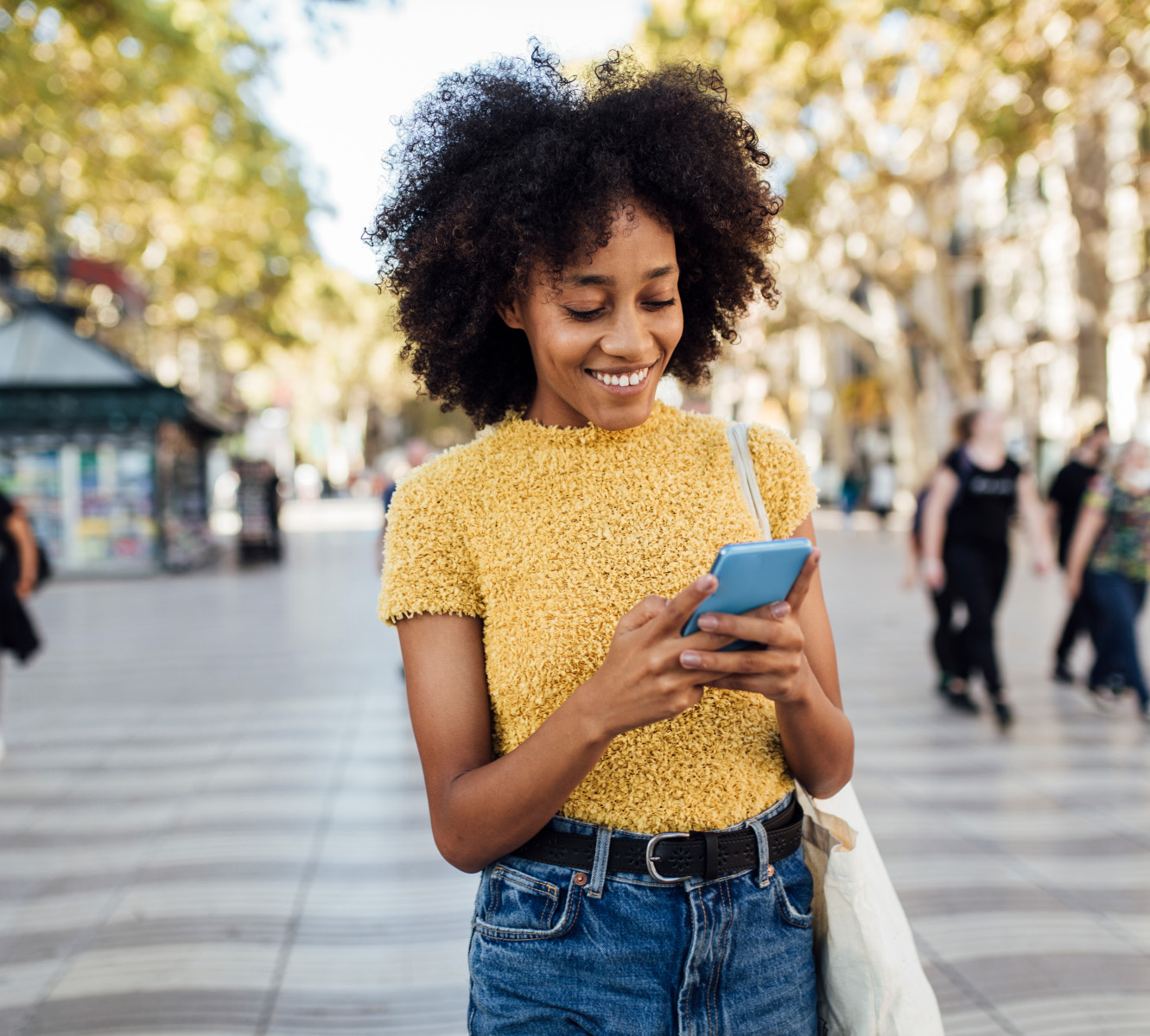 Ung kvinde iført gul bluse og jeans, står midt på et torv og smiler ad hendes telefon. 